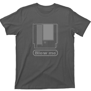 May include: A dark gray t-shirt with a white outline of a video game cartridge and the text "Blow me" printed on the front.