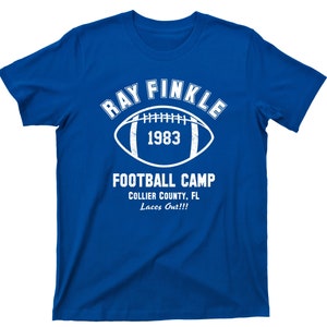May include: A royal blue t-shirt with a white graphic design. The graphic features a football with the text "Ray Finkle 1983 Football Camp Collier County, FL Laces Out!!!"