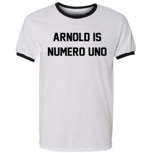 May include: White t-shirt with a black ringer and the text "ARNOLD IS NUMERO UNO" printed in black.