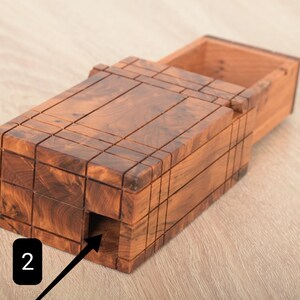 May include: A rectangular wooden puzzle box with a rich brown grain pattern and a sliding lid mechanism. The exterior features a grid of carved lines and a small square opening on the side marked with a black square containing the white number 2.