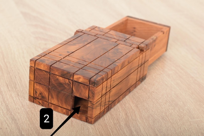 May include: A rectangular wooden puzzle box with a rich brown grain pattern and a sliding lid mechanism. The exterior features a grid of carved lines and a small square opening on the side marked with a black square containing the white number 2.