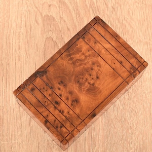 Peut inclure: Une boîte rectangulaire en bois avec un motif de grain de loupe brun riche. La boîte présente une série de lignes parallèles gravées sur la surface, créant un aspect texturé. La boîte est posée sur une surface en bois de couleur claire.