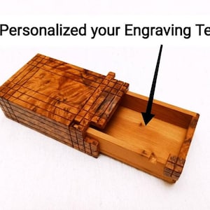 May include: A rectangular wooden box with a sliding drawer mechanism. The exterior features a dark brown wood grain with a grid-like pattern. The interior is a lighter natural wood tone. Text above reads: Personalized your Engraving Text.