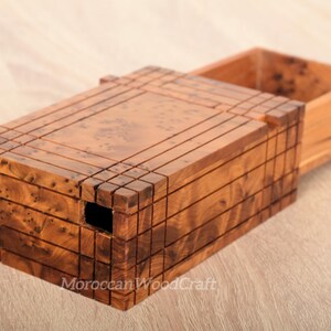 以下が含まれることがあります： 複雑な模様のついた木製の箱で、スライド式の蓋が付いています。箱が開いており、内側が見えています。箱は明るい茶色の木でできており、濃い茶色の木目が付いています。