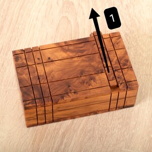 May include: A rectangular wooden puzzle box featuring a natural brown wood grain pattern with multiple horizontal and vertical grooves. A black arrow graphic points upward next to a black rectangular icon containing the white number 1.