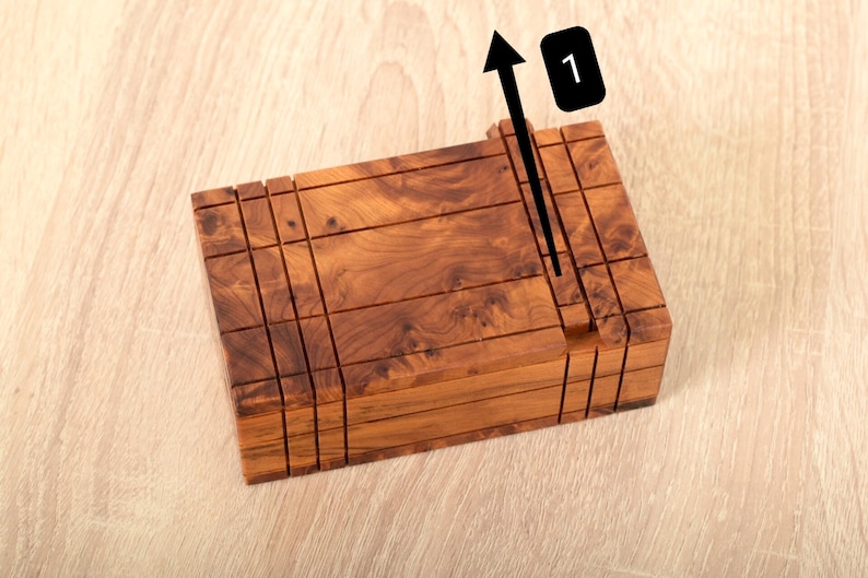 May include: A rectangular wooden puzzle box featuring a natural brown wood grain pattern with multiple horizontal and vertical grooves. A black arrow graphic points upward next to a black rectangular icon containing the white number 1.