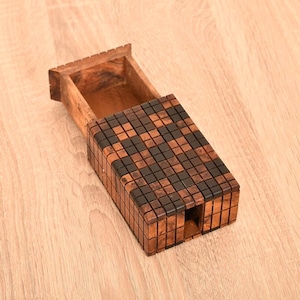 Peut inclure: Une boîte en bois avec un tiroir coulissant, présentant un motif en mosaïque de carrés marron foncé et clair. La boîte est ouverte, révélant l'intérieur, et repose sur une surface en bois de couleur claire.