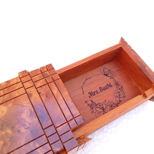 May include: A rectangular wooden box with a sliding lid featuring a natural burl wood grain pattern. The interior base of the box is engraved with a floral wreath design surrounding the text Mrs. Sushi, centered within the open compartment.