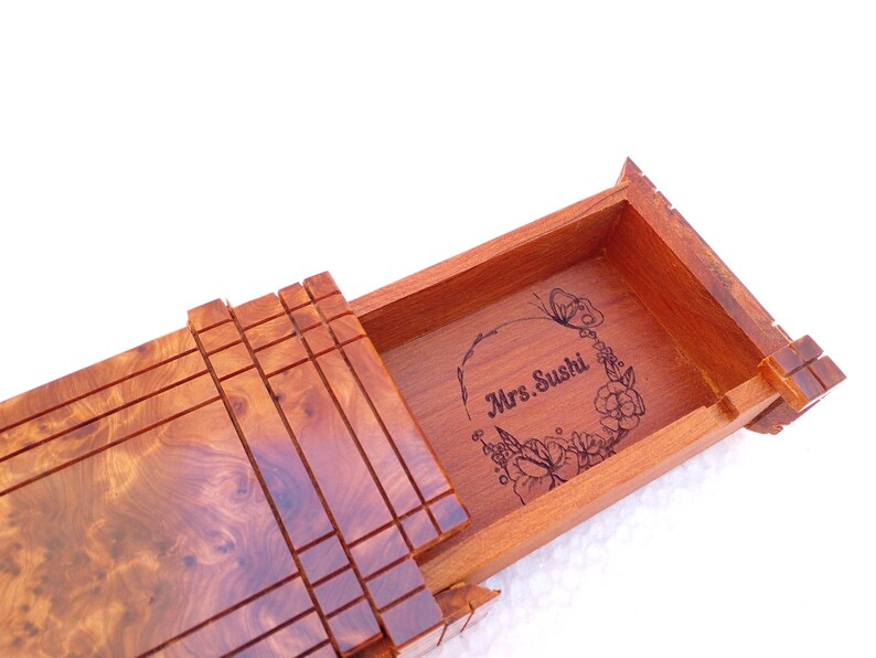 May include: A rectangular wooden box with a sliding lid featuring a natural burl wood grain pattern. The interior base of the box is engraved with a floral wreath design surrounding the text Mrs. Sushi, centered within the open compartment.