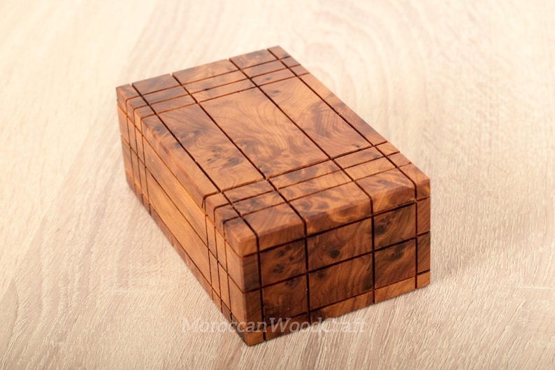 May include: A rectangular wooden box featuring a natural brown burl wood grain pattern. The exterior surface is divided into a grid of small, recessed square and rectangular sections, creating a textured, geometric appearance across the top and sides.
