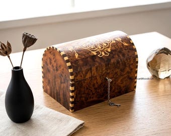 May include: A decorative wooden chest with a curved top, featuring a dark brown finish with lighter wood accents and a gold-colored inlay design. The chest has a small key hanging from a chain. A black vase with dried flowers and a crystal sphere are also present.