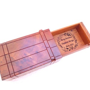 May include: A rectangular wooden box with a sliding lid featuring a dark reddish-brown grain. The interior base is engraved with a circular wreath design surrounding the text: Merry Christmas, Chet Love Always, Mom 2022.