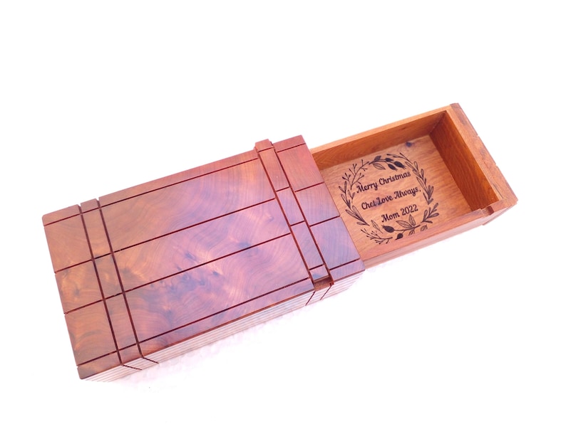 May include: A rectangular wooden box with a sliding lid featuring a dark reddish-brown grain. The interior base is engraved with a circular wreath design surrounding the text: Merry Christmas, Chet Love Always, Mom 2022.
