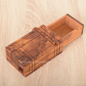May include: A rectangular wooden box with a sliding lid mechanism, crafted from brown wood with a visible natural grain pattern. The exterior features a grid of etched lines, and the lid is partially open, revealing a hollow interior compartment.