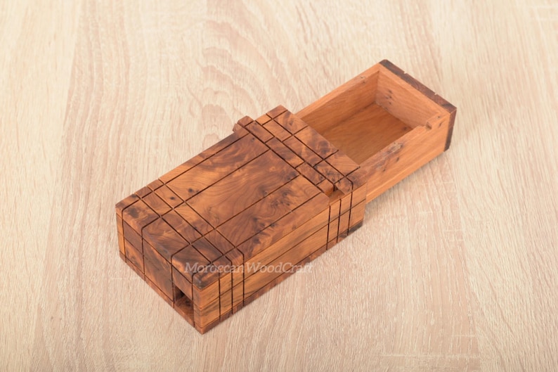 May include: A rectangular wooden box with a sliding lid mechanism, crafted from brown wood with a visible natural grain pattern. The exterior features a grid of etched lines, and the lid is partially open, revealing a hollow interior compartment.