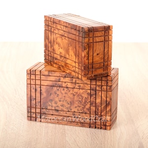 Può includere: Due scatole di legno impilate l'una sull'altra. Le scatole sono in legno marrone chiaro con intarsi in legno marrone scuro. Le scatole hanno un motivo geometrico sul davanti e sui lati. Le scatole sono appoggiate su una superficie in legno marrone chiaro.