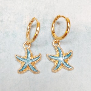 May include: A pair of gold-coloured hoop earrings featuring starfish charms. The starfish are light blue with gold-coloured outlines and details. The earrings are displayed on a light blue and white surface.