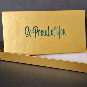 May include: A gold gift box with a white interior. The lid of the box has the words "So Proud of You" printed in green.