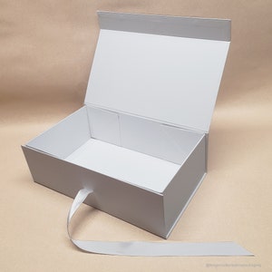 May include: White gift box with a satin ribbon. The box is open and the inside is empty. The box is made of sturdy cardboard and has a smooth finish.