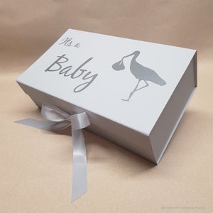 May include: White gift box with a silver ribbon and a silver stork design. The box has the text "It's a Baby" printed on the top.