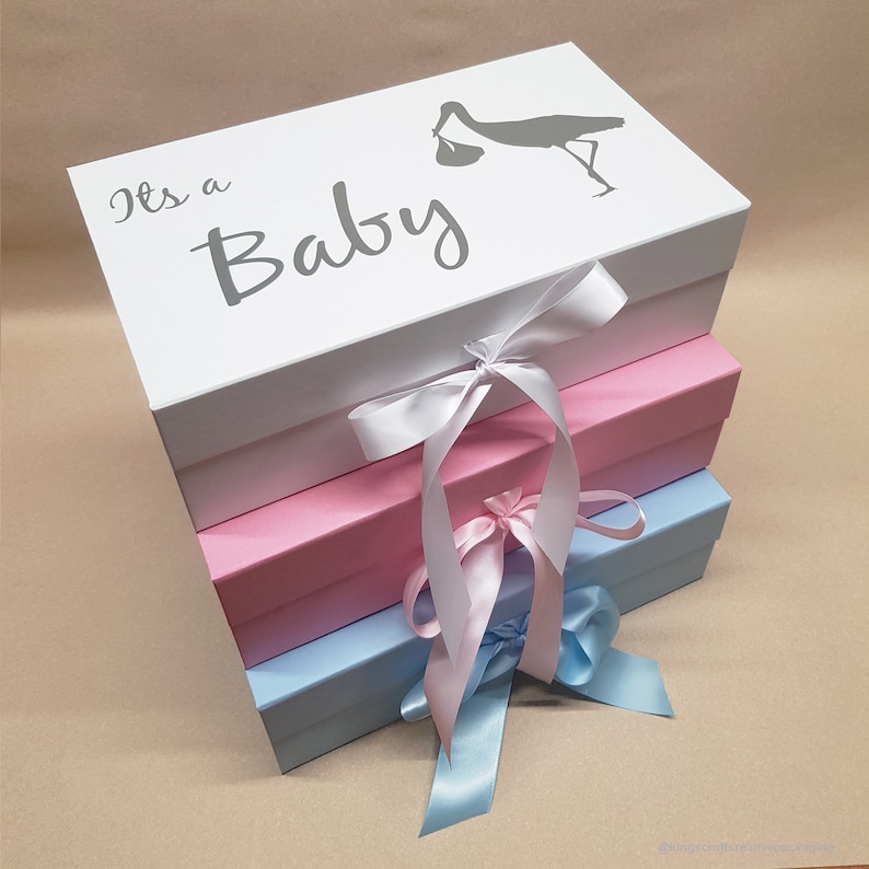 May include: A stack of three boxes with white, pink, and blue lids. The top box has a white background with the text "It's a Baby" and a gray silhouette of a stork carrying a baby. Each box has a satin ribbon tied around it.