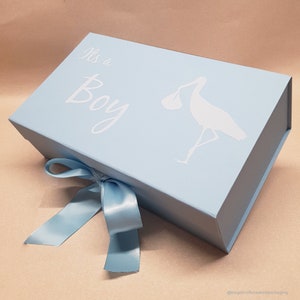May include: A light blue gift box with a white ribbon. The box has a white stork silhouette carrying a baby and the text "It's a Boy" on the top.