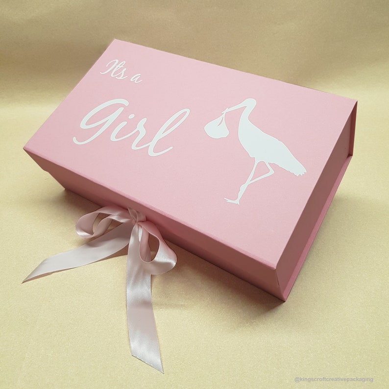 May include: Pink gift box with a white ribbon and a white stork carrying a baby. The box has the text "It's a Girl" on the front.