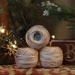 May include: Three balls of beige and white embroidery floss, stacked on top of each other. The label on the top ball reads "Valdani 8 Colorfast JP 4".