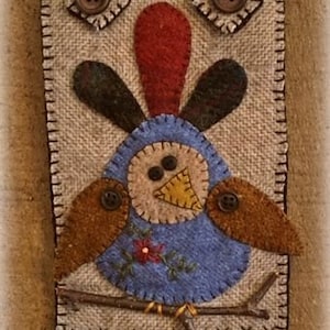 May include: A brown, blue, and red felt bird applique with a twig and buttons. The bird is perched on a twig and has a red flower embroidered on its chest. The applique is attached to a brown burlap background.