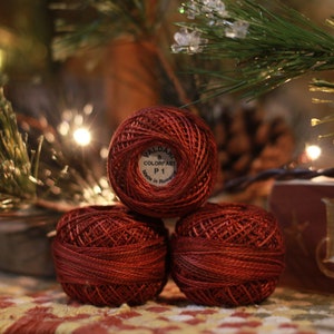 May include: Three spools of red embroidery thread are stacked in a pyramid shape. The top spool has a label that reads "VALDANI COLORFART P1". The background includes Christmas tree branches, lights, and a wooden sign.