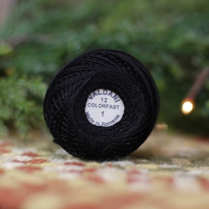 May include: A black Valdani 12 Colorfast thread ball with a white label that reads "1" and "Made in Romania". The thread ball is on a patterned surface with a blurred green background.