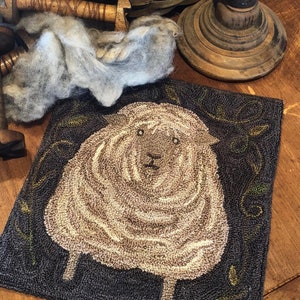 May include: A square, hand-stitched rug featuring a sheep design. The sheep is depicted in shades of tan and cream against a dark blue background. Green leaf accents border the sheep. The rug is placed on a wooden surface.
