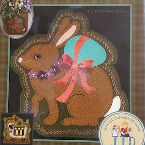 May include: A brown felt applique of a bunny with a teal egg and a pink bow. The design is 9 x 10 inches and includes the text "Special Delivery". The image is a seasonal ornament.