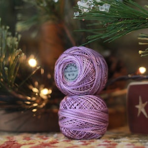 May include: Two spools of lavender-colored embroidery floss, one on top of the other. The top spool has a white label with the text "Valdani Colorfast M 118".
