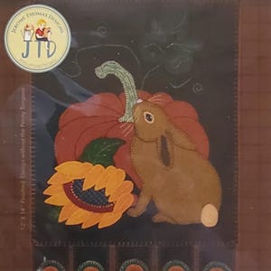 May include: A wool applique kit titled "Pumpkin Eater" featuring a brown rabbit, a large orange pumpkin, and a sunflower. The design is on a dark background. The kit is 12" x 14" and includes instructions.