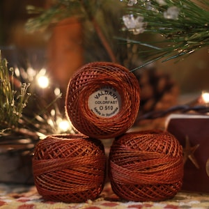 May include: Three spools of Valdani 8 Colorfast thread in a warm, reddish-brown hue. The top spool displays the brand name and color details. The spools are arranged in a triangular stack, with a blurred background of Christmas decorations.