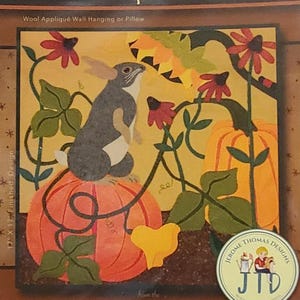 May include: A wool appliqué pattern titled "The Pumpkin Patch" featuring a gray bunny sitting on an orange pumpkin. The design includes red flowers, green leaves, and the text "Bunny Rabbit Series - AUTUMN". The finished design is 17" x 17".
