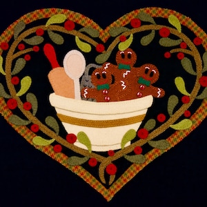 May include: A heart-shaped wreath with green leaves and red berries surrounds a white bowl filled with gingerbread men. The bowl also contains a wooden rolling pin and a wooden spoon.