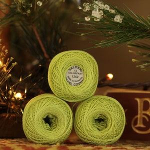 May include: Three balls of lime green Valdani 12 Colorfast Pearl Cotton thread are stacked in a pyramid shape. The thread is labeled with the number 1262. The background includes a Christmas tree and a wooden sign.