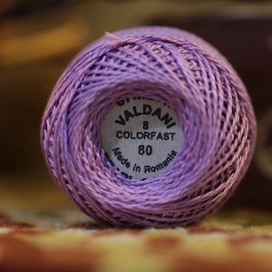 May include: A close-up of a ball of lavender-colored thread. The thread is labeled "VALDANI COLORFAST 80" and "Made in Romania". The thread is tightly wound, showing the texture of the fibers. The background is blurred.