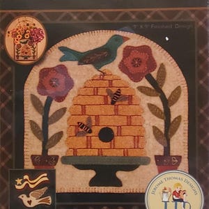 May include: A seasonal wall pocket design titled "The Bird and the Bees." The design features a bird perched atop a brick beehive, surrounded by flowers and bees. The text "The Bird and the Bees" is displayed at the top. The finished design is 9 x 9 inches.