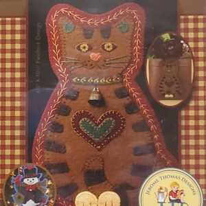 May include: A brown and red cat-shaped wall pocket with the text "Love Mew". The cat has a heart, bell, and decorative stitching. Includes a snowman pin and a Jerome Thomas Designs logo.