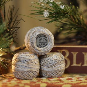 May include: Three spools of white and silver embroidery floss, stacked on top of each other. The top spool has a label that reads "67 meter Valdani 8 Colorfast M 23 Pearl Made in Holland".