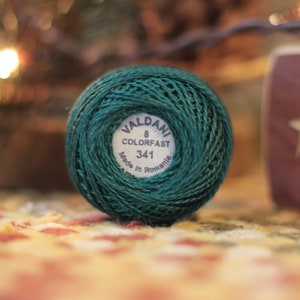 May include: A ball of Valdani 8 Colorfast thread in a deep teal green. The label reads "341 Made in Romania." The thread is tightly wound, showcasing its texture. The background is blurred, with hints of a Christmas tree and a decorative item.
