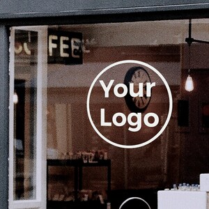 May include: A white circle with the text "Your Logo" in black lettering on a storefront window. The window is behind a black frame and there are lights hanging from the ceiling.