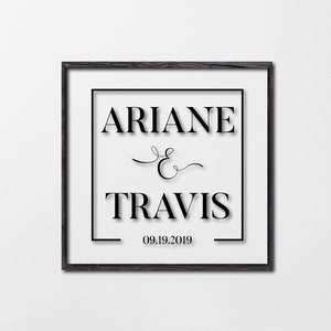 May include: A framed print with the names "ARIANE & TRAVIS" in a bold, elegant font, with the date "09.19.2019" below. The print is set against a white background, framed in a dark wood.