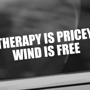 May include: A white sticker on a car window with the text "Therapy is pricey, wind is free."