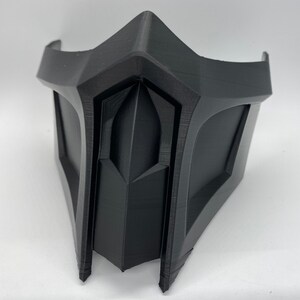 Puede incluir: Una m&aacute;scara de pl&aacute;stico negro con un dise&ntilde;o geom&eacute;trico. La m&aacute;scara tiene una forma afilada y angular con una barbilla puntiaguda y una secci&oacute;n triangular elevada en el centro.