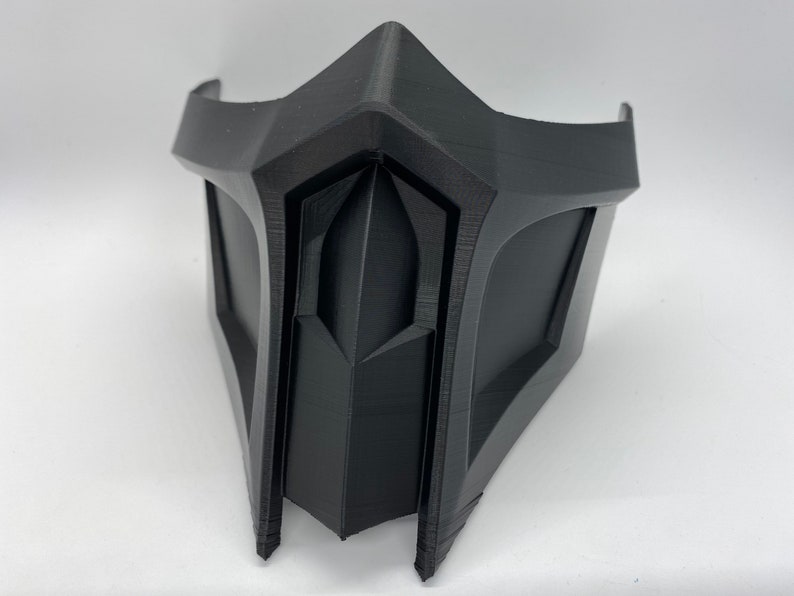 Puede incluir: Una m&aacute;scara de pl&aacute;stico negro con un dise&ntilde;o geom&eacute;trico. La m&aacute;scara tiene una forma afilada y angular con una barbilla puntiaguda y una secci&oacute;n triangular elevada en el centro.