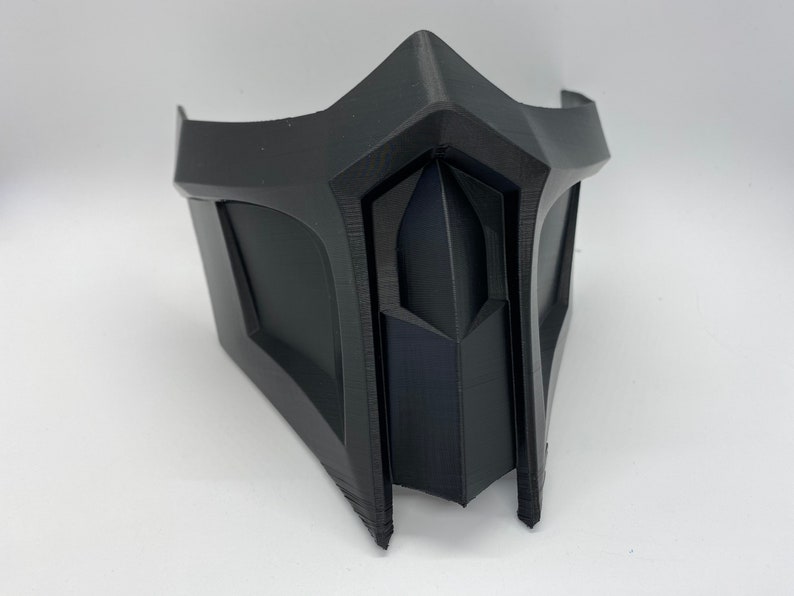 Puede incluir: Una m&aacute;scara de pl&aacute;stico negro con un dise&ntilde;o geom&eacute;trico. La m&aacute;scara tiene una parte superior puntiaguda y una parte inferior ancha y plana. Tiene una peque&ntilde;a abertura rectangular en el centro.